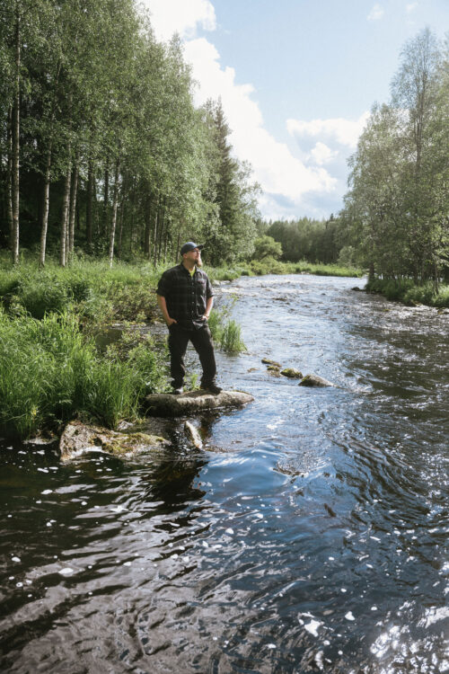 Jaakko Alasirniö seisoo virtaavan veden törmällä kiven päällä, takana koivumetsä.